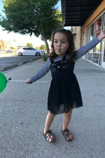 Child in a black cat dress holding a green balloon, sticking out her tongue playfully.
