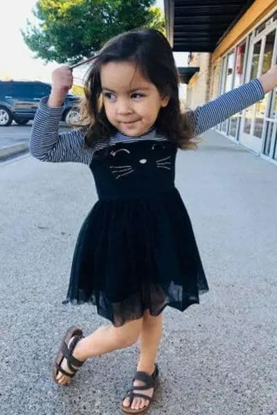 Child in a black dress with cat design, smiling and posing outside a building.